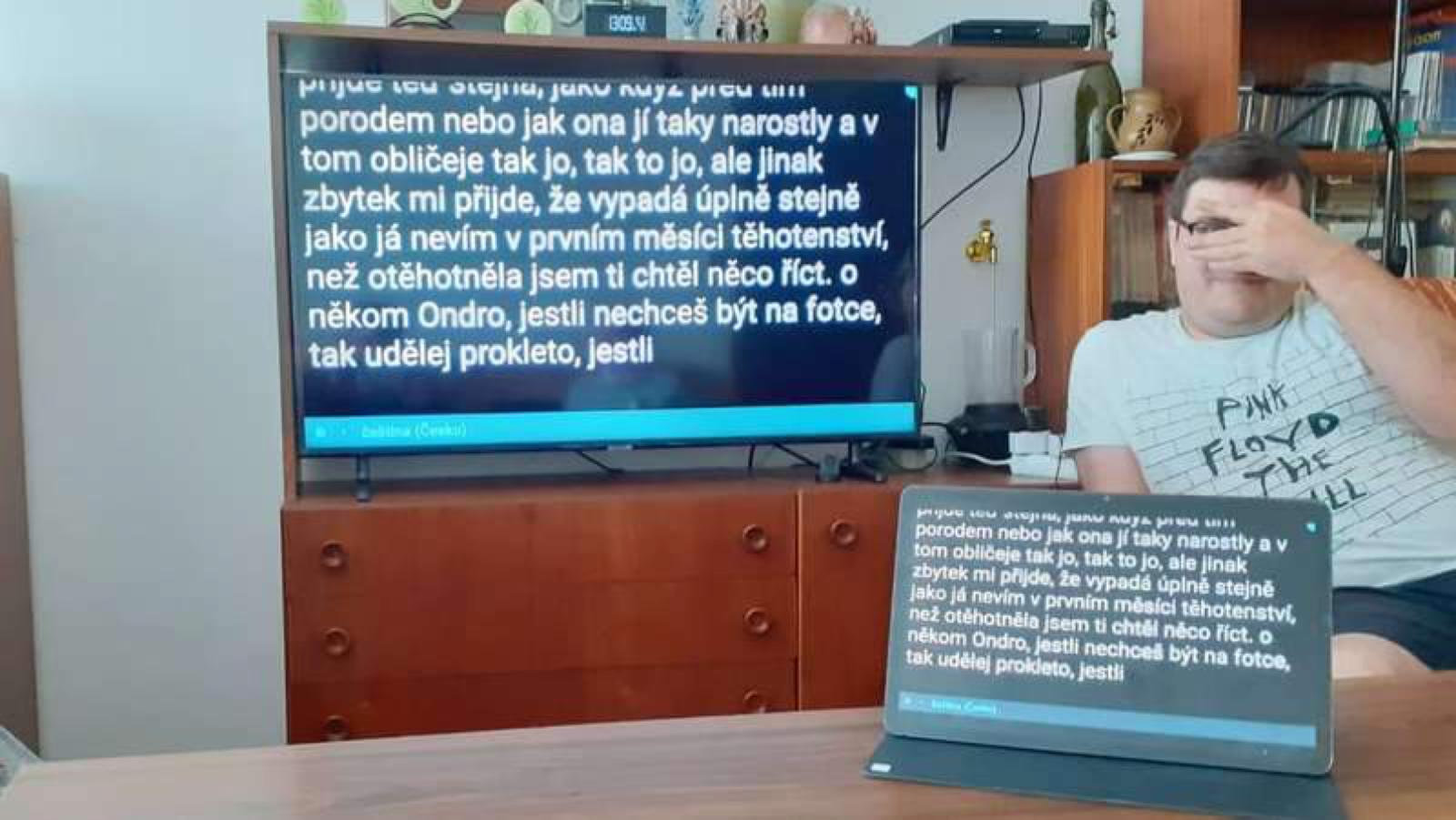 na stole je tablet, kde je spuštěna aplikace na rozpoznání mluvy a její převod do textové formy a obrazovka tabletu se promítá na televizor.